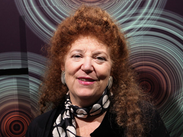 A white woman with wavy red hair, with dark earings and a neckscarf, standing against a backdrop of concentric circles